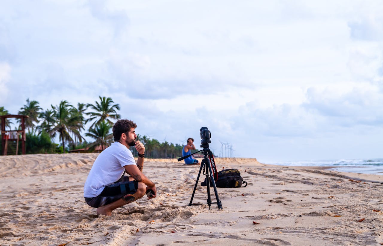 Services A photographer kneels by a camera on a sandy beach, capturing scenic views in Barra de Camaratuba, Brazil.